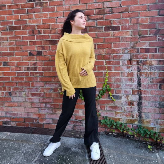 A person wearing an off-shoulder yellow jumper with a wide neckline, paired with black pants and white shoes, standing against a brick wall background.