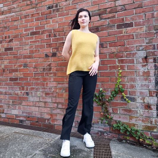 A person wearing a yellow sleeveless tank top with ribbed details on the sides, paired with black trousers and white shoes, standing against a brick wall background.