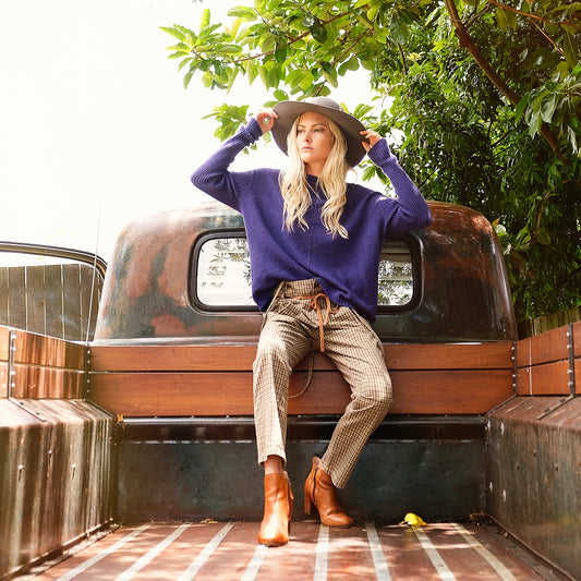 Blond lady sitting on the back of a truck holding her hat. She is wearing a purple cashmere possum silk jumper made in new zealand that has angled rib details on the body and ribbed long sleeves