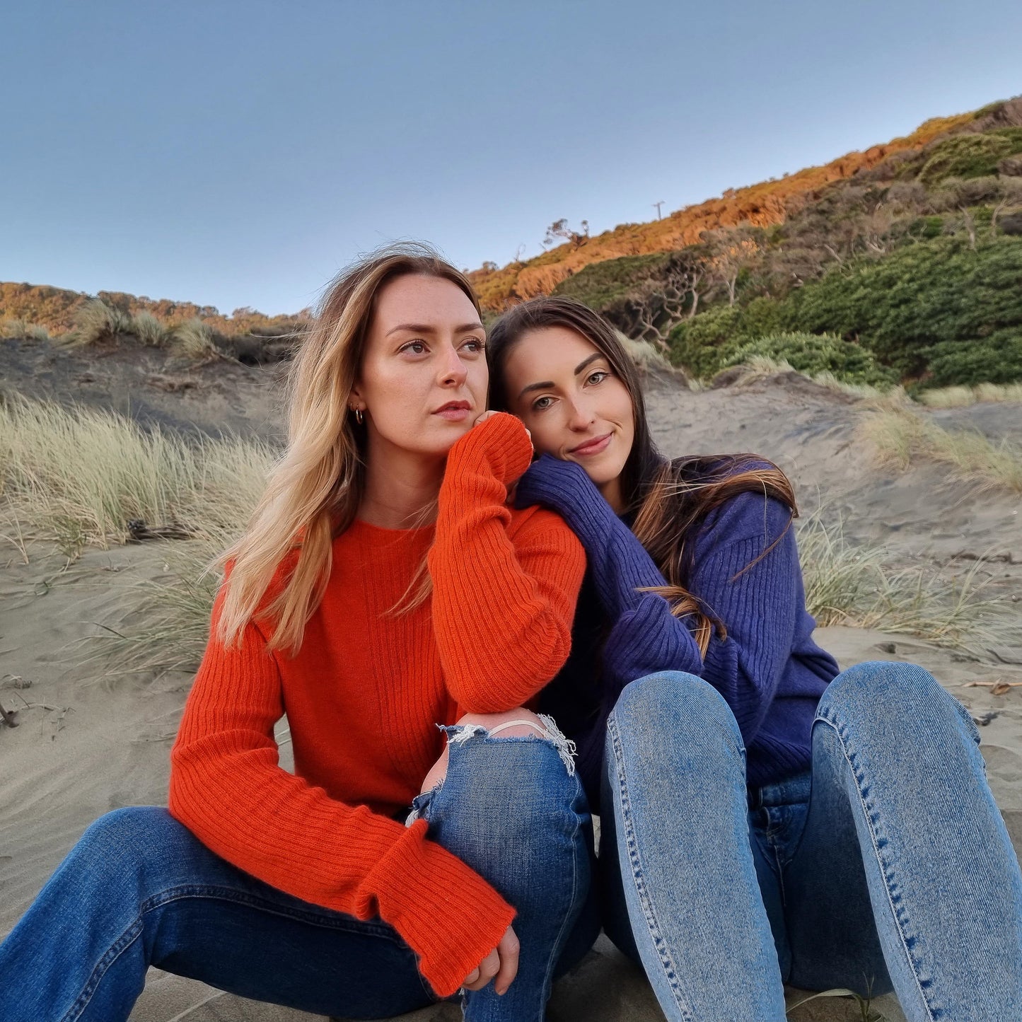 Two friends sitting cosy together on the beach, both are wearing cashmere possum silk jumpers that were made in new zealand, the blond girl is wearing a bright orange jumper and the brunette girl is wearing a purple sweater.