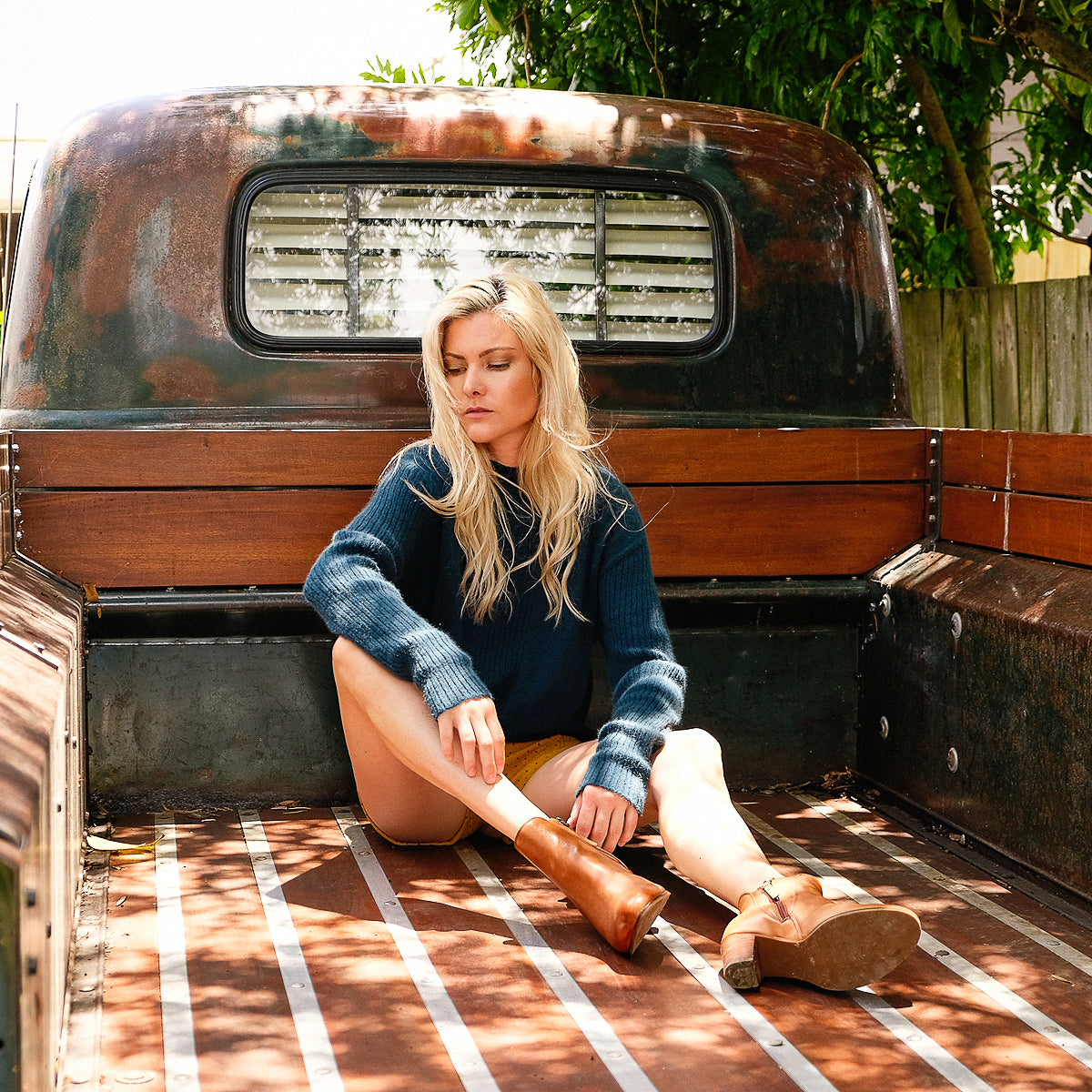 Blond lady sitting on the back of a truck. She is wearing a teal blue cashmere possum silk jumper made in new zealand that has rib detailing on the centre front of the jumper body and ribbed long sleeves