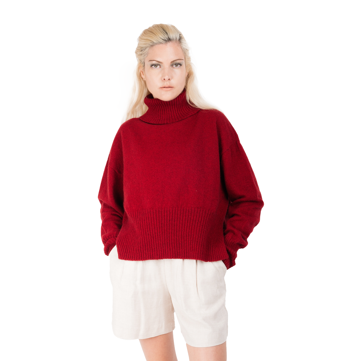 Blond lady wearing a bright red jumper. Cropped body with a long split ribbed hem. Turtleneck and ribbed cuffs. Sweater made from Merino possum and nylon and made in new zealand. She has her hands in her pockets and is looking forward
