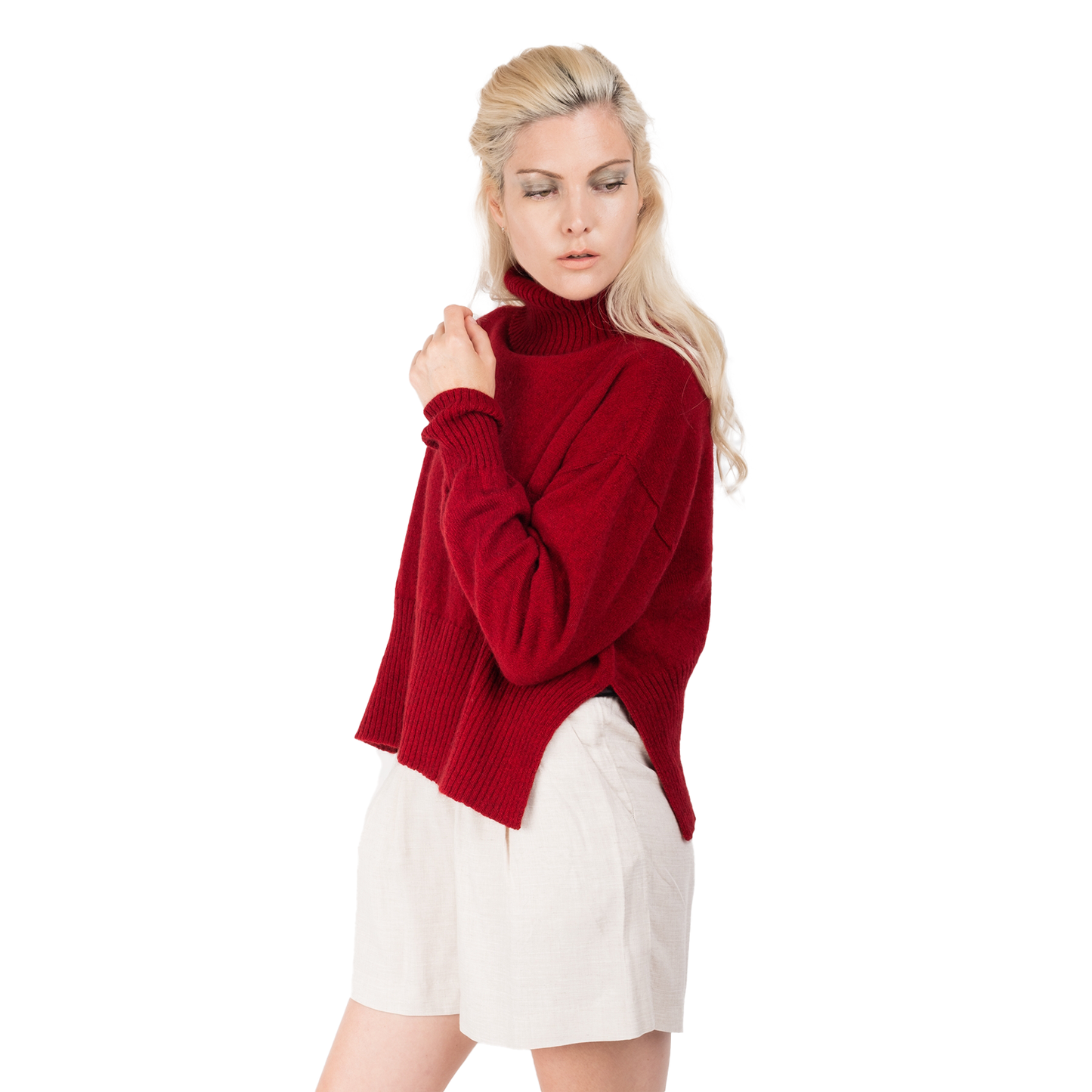 Blond lady wearing a bright red knitted jumper. Cropped body with a long split ribbed hem. Turtleneck and ribbed cuffs. Sweater made from Merino possum and nylon and made in new zealand. She is facing the side with her arm up to her shoulder showing off the split in the sides of the sweater