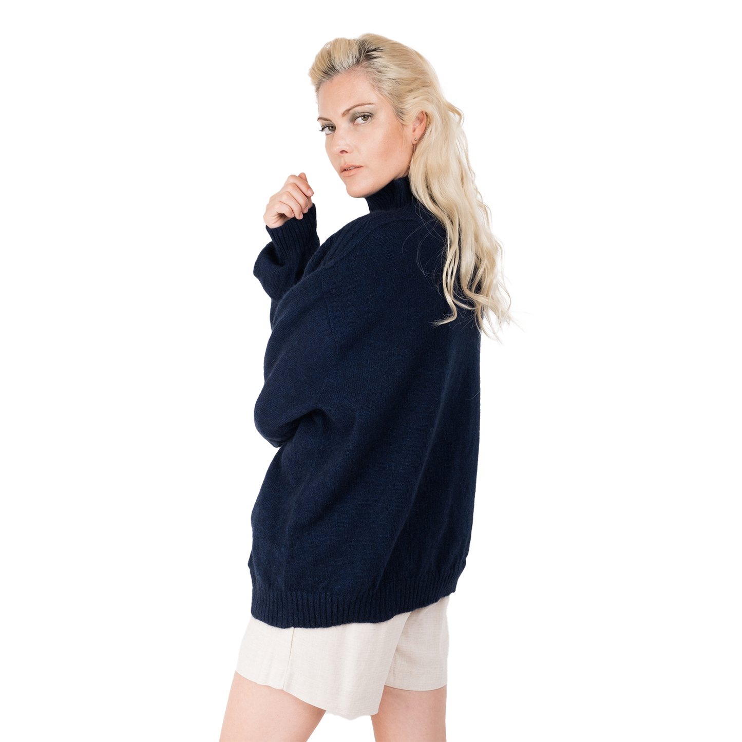 Blond lady wearing a deep inky blue knitted jumper. Full oversizebody with a  ribbed hem. Turtleneck and ribbed cuffs. Sweater made from Merino possum and nylon and made in new zealand. She is facing  slighty to the back with her arm up near her face showing off the ribbed line detail on the top of the sleeves of the sweater as well as the back